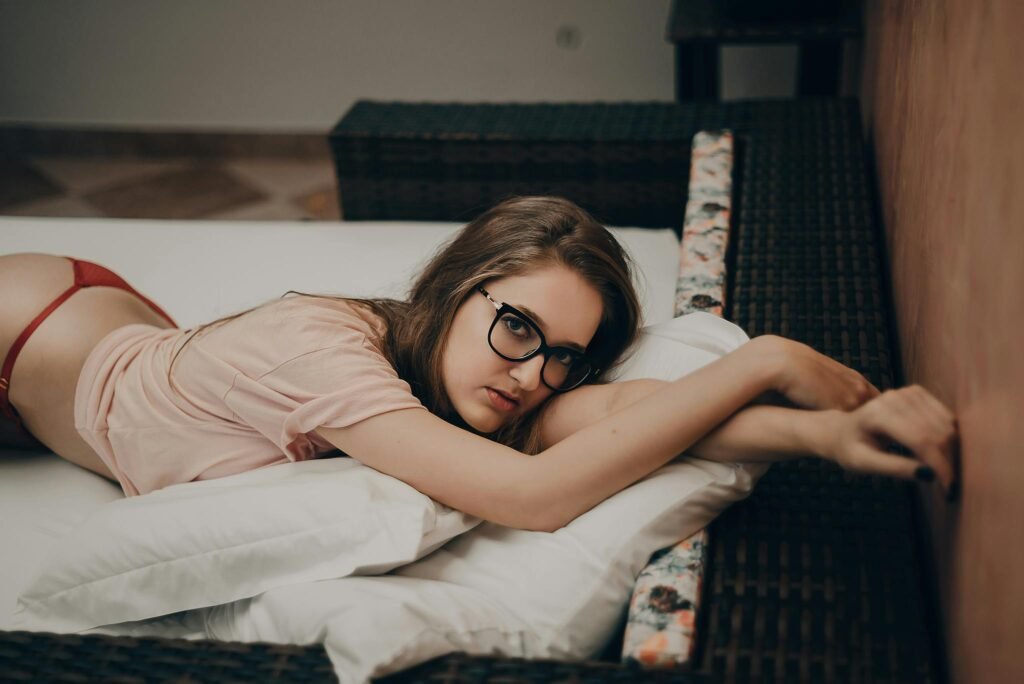 Young woman relaxing on a bed indoors, wearing glasses and casual attire.