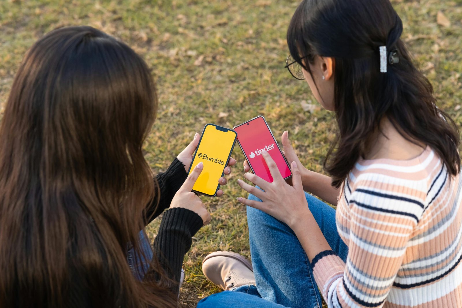 Two women using dating apps Bumble and Tinder on smartphones in a park setting.