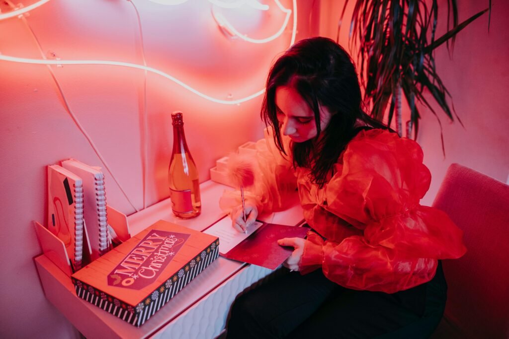 A woman writes a holiday card in a vibrant, neon-lit room with a festive atmosphere.