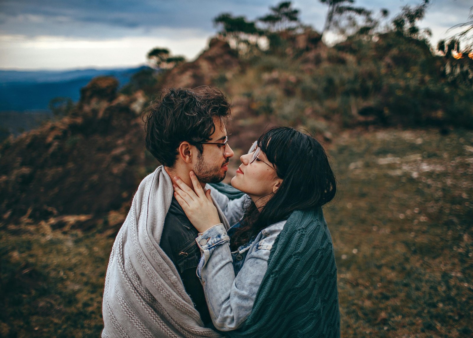 A young couple embraces warmly under a blanket in a natural outdoor setting.