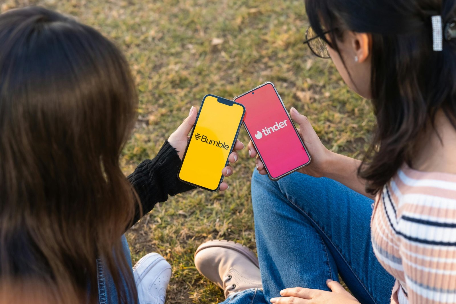 Two women using Bumble and Tinder apps on smartphones in a park setting.