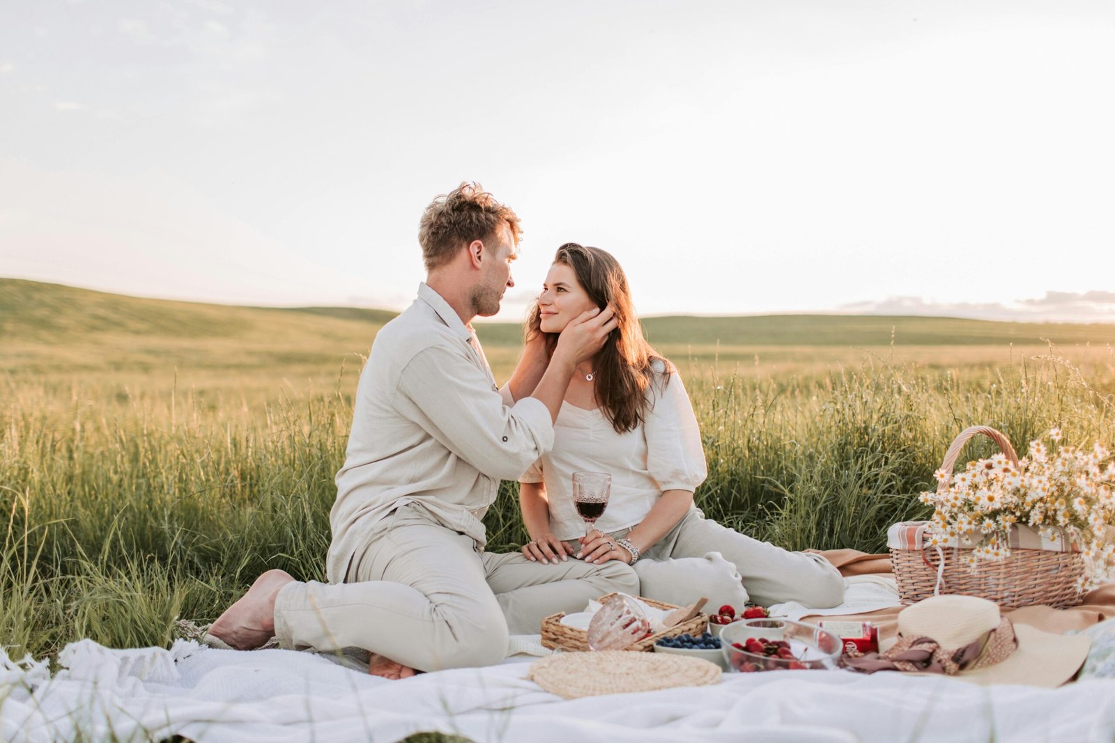 A couple enjoying a romantic picnic at sunset in a grassy field, surrounded by nature.